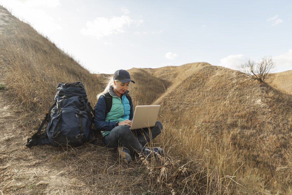 Woman sitting on a grassy hillside with a backpack, studying on a laptop outdoors