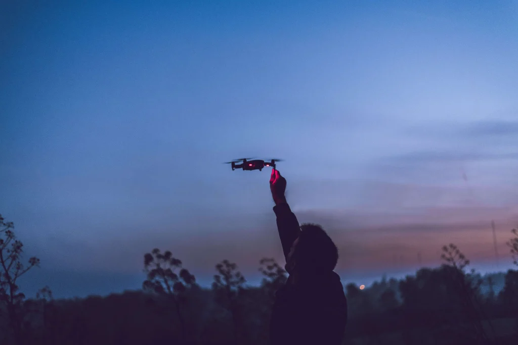 Drone pilot silhouetted at dusk reaching up to hand-catch a small drone showing red navigation lights